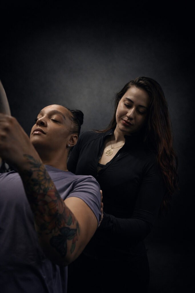 A trainer stands closely behind a client during an exercise, providing gentle hands-on guidance as the client moves with eyes closed, conveying trust, focus, and attentive coaching in a dark, studio-lit setting.