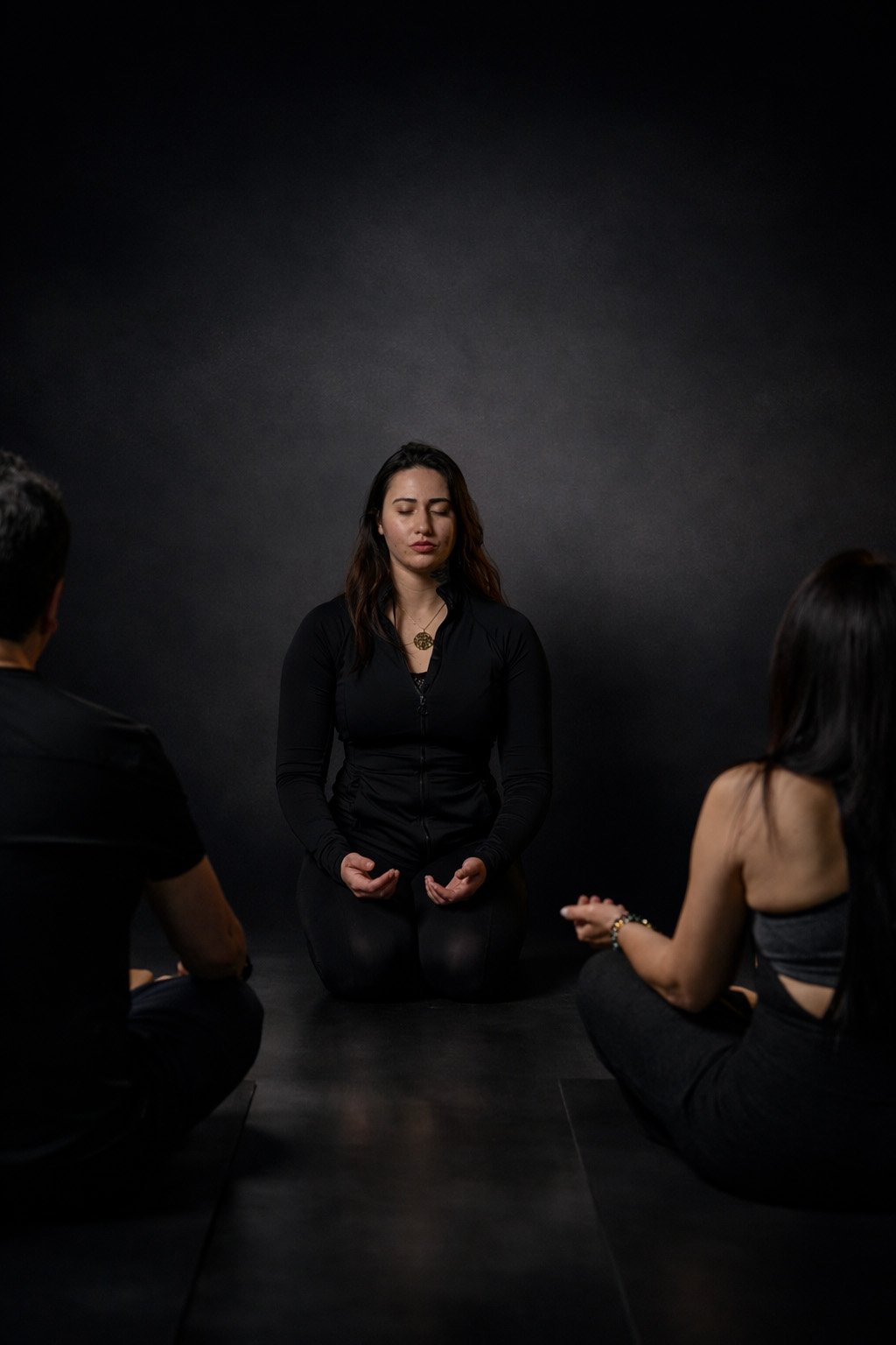 A trainer sits calmly with eyes closed and hands resting open on her knees, centered between two seated clients, suggesting a guided moment of stillness, breathwork, or nervous system regulation in a dark, studio-lit space.