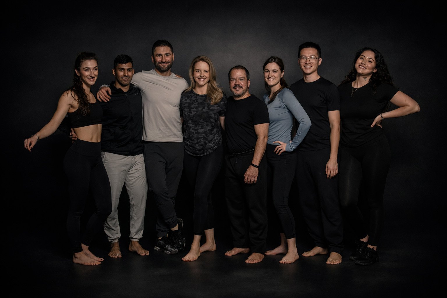 A diverse group of trainers and clients stand together barefoot in a relaxed line against a dark, studio-lit background, smiling and leaning into one another, conveying community, trust, and shared commitment to training.