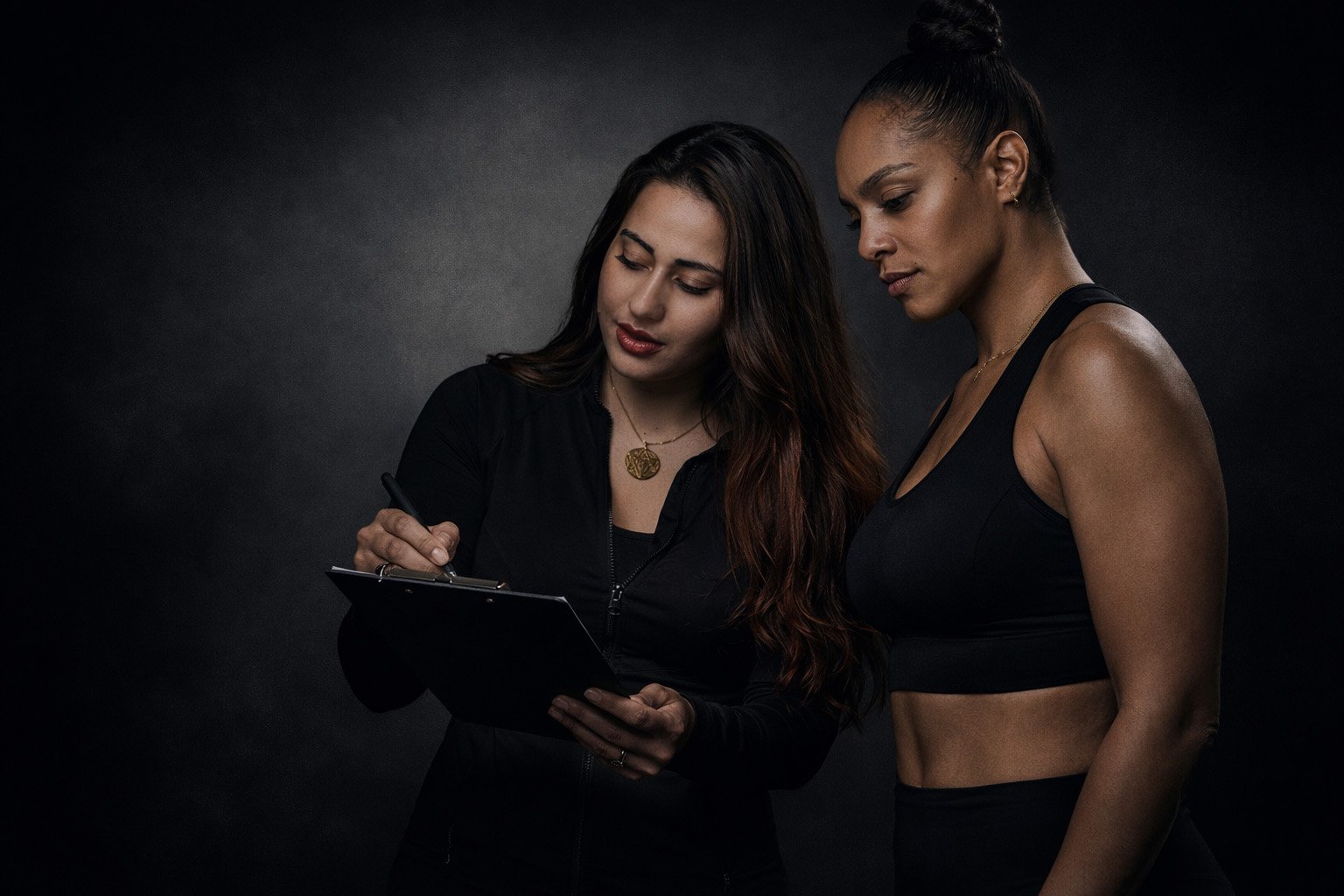 A trainer and a client stand side by side in a dark, studio-lit setting, reviewing notes on a clipboard as the trainer writes while the client looks on attentively, suggesting a personalized fitness assessment or planning session.