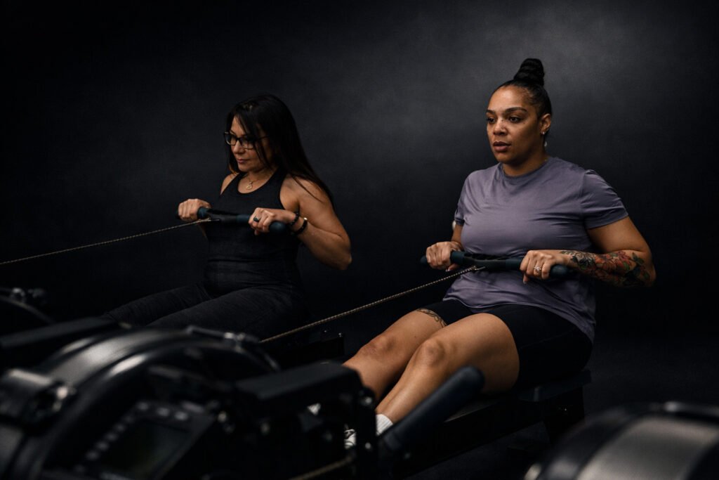 Two women exercise side by side on rowing machines in a dark, studio-style gym, pulling the handles toward their torsos with focused expressions, emphasizing strength, effort, and controlled movement.