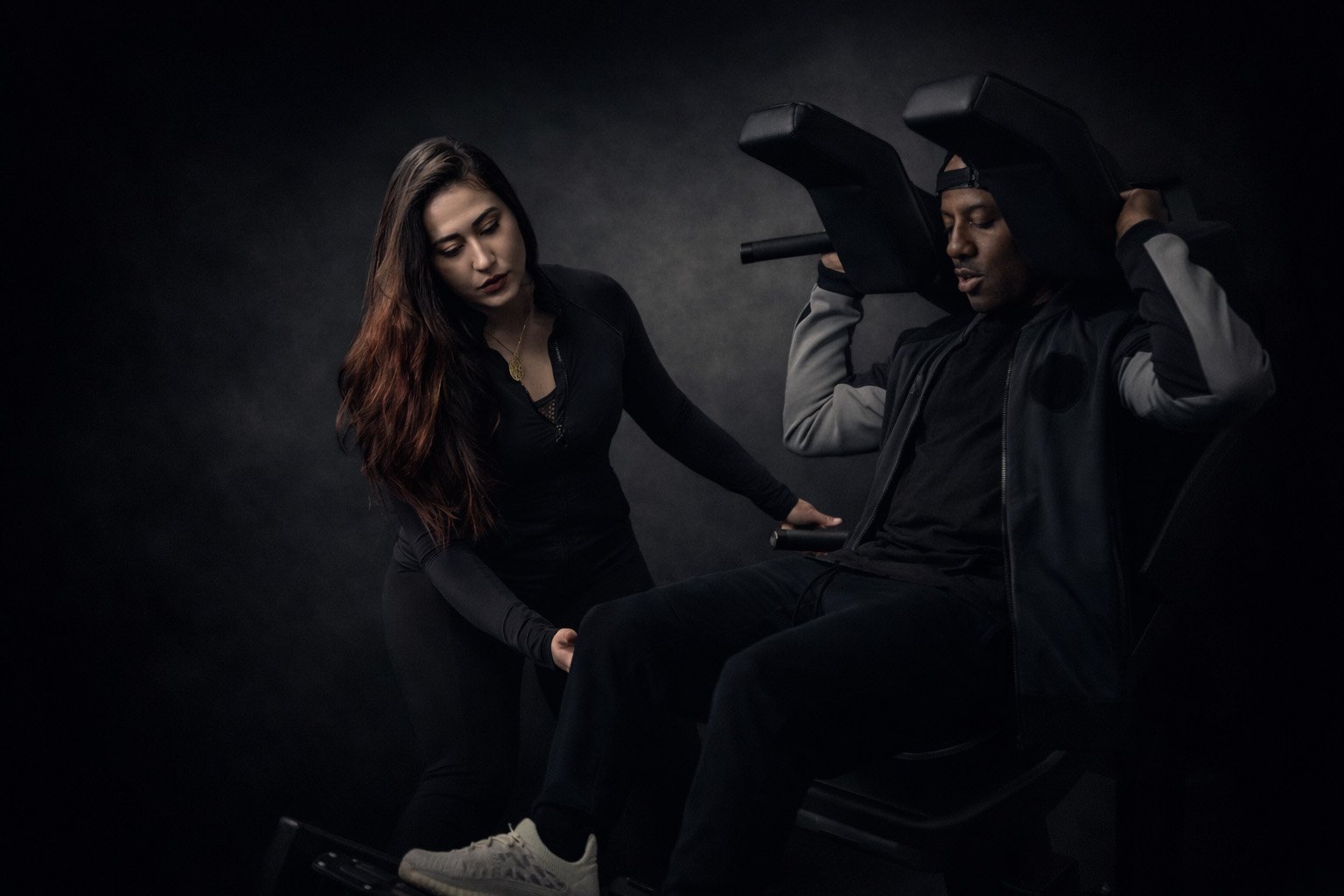 A personal trainer stands beside a seated exerciser using a weight machine, guiding their leg position with one hand while the person presses the padded handles overhead, set against a dark, studio-style gym background.