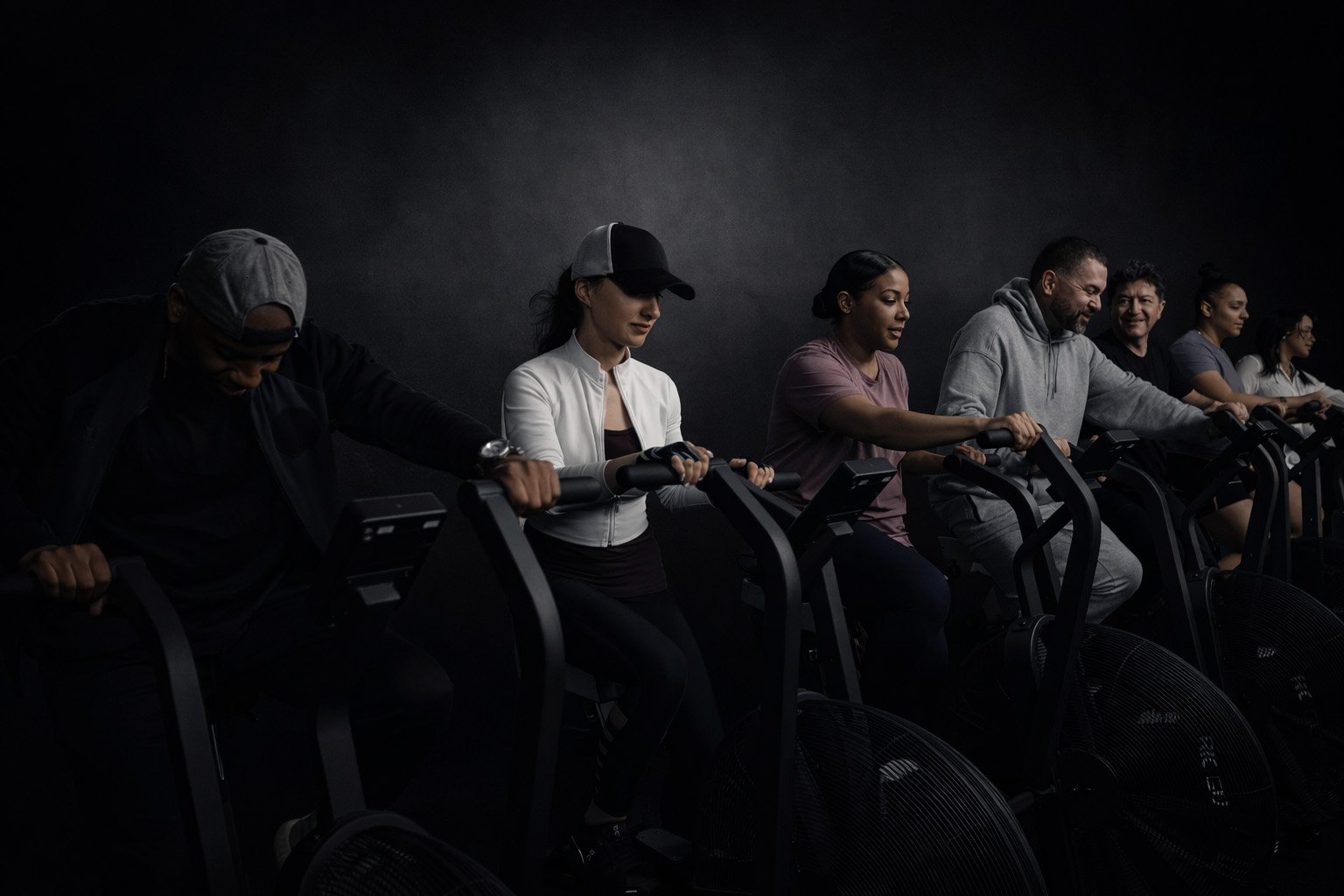 A diverse group of people exercise side by side on air bikes in a dark, studio-style gym, leaning forward as they pedal and push the moving handles, conveying intensity, teamwork, and shared effort.